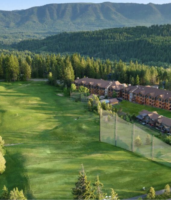 An aerial view showcases the lush green golf course of Suncadia Resort, nestled among adjacent buildings. It is beautifully surrounded by dense forests and majestic mountains in the background.